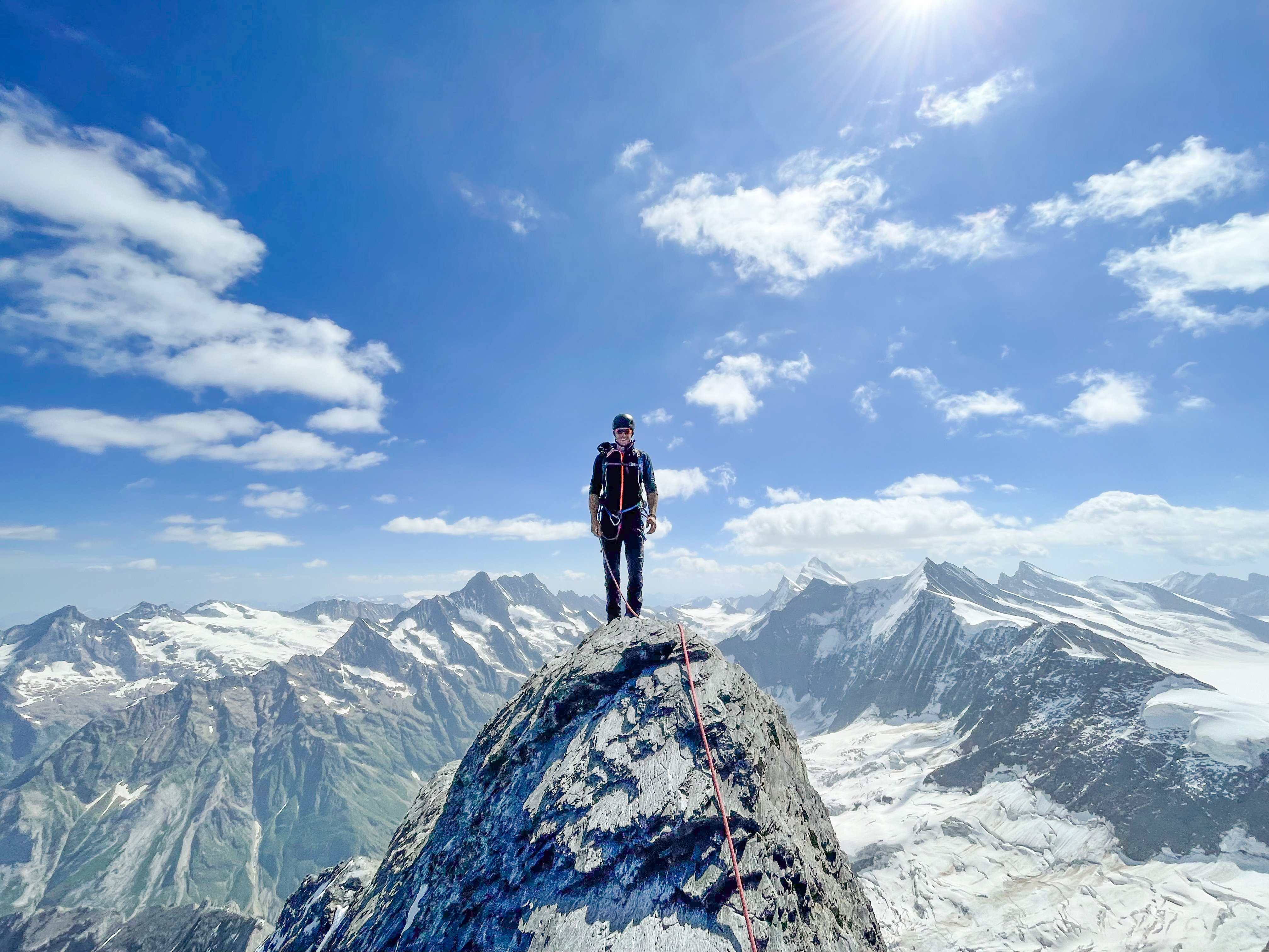 Person auf Berggipfel mit Kletterausrüstung und Seil, umgeben von verschneiten Alpen.